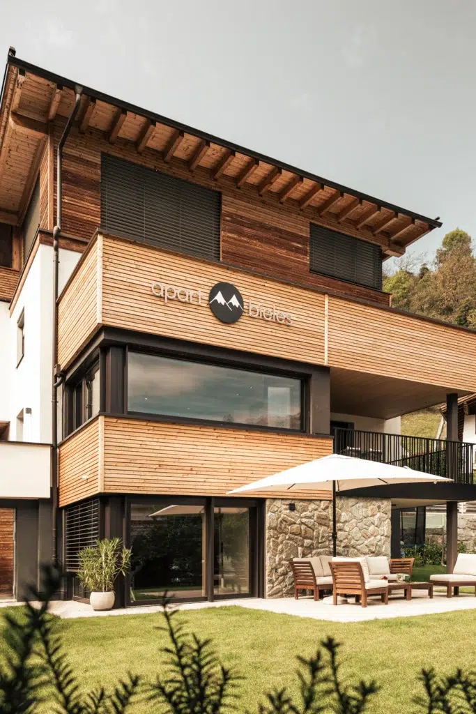 Modernes hölzernes Apart Bieles-Gebäude mit Sitzgelegenheiten im Freien, großen Fenstern und einem Sonnenschirm im Innenhof.